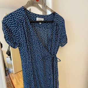Blå blommig klänning från & Other Stories -  Blå klänning med små vita blommor från & Other Stories. Klänningen har en omlott-design med knytband i midjan och korta ärmar. 💗 Liten i storleken! 