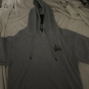 Grå hoodie från Stussy - Säljer en grå hoodie från Stussy med deras ikoniska logga tryckt både fram och bak. Hoodien har en justerbar huva med snören och korta ärmar. Perfekt för en avslappnad stil. Om ni har fler frågor är det bara att skriva.