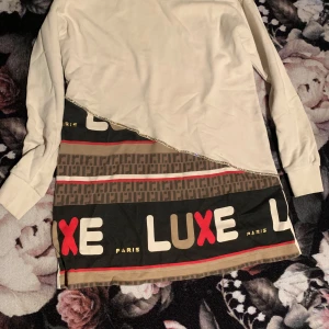 Vit och mönstrad långärmad tröja med text - Säljer en unik långärmad tröja i vitt med en mönstrad nederdel. Tröjan har texten 'LUXE' i stora bokstäver och detaljer i rött, svart och beige. Perfekt för dig som vill sticka ut med en trendig stil.