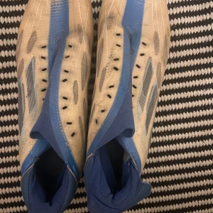 Blå och vita fotbollsskor - Säljer ett par blå och vita fotbollsskor med dobbar. Skorna har en strömlinjeformad design med blå detaljer och är perfekta för spel på gräsplaner.