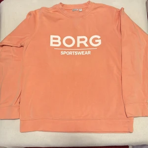Persikofärgad tröja från Björn Borg - I enkelheten vardag eller när man tränar. Inte så mycket använd. Säljer en snygg persikofärgad tröja från Björn Borg med texten 'BORG SPORTSWEAR' i vitt på framsidan. Tröjan har långa ärmar och en rund halsringning. Perfekt för en avslappnad stil.