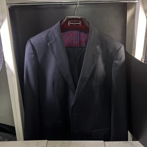 Skräddarsydd Mörkblå randig kostym från Giorgio Vallino - Elegant mörkblå kostym från Giorgio Vallino med subtila ränder. Kavajen har en klassisk passform med två knappar och ett färgglatt foder i lila och rött mönster. Perfekt för formella tillfällen, kommer göra dig den mest välklädda på plats. Eftersom detta är valt efter personliga efterfrågningar så är den mycket speciell. Hör av er vid minsta fundering🙌