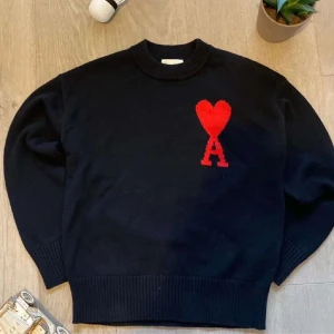 Ami Paris Sweater  - Snygg mörkblå tröja med ett rött hjärta och bokstaven 'A' på bröstet. Tröjan har en ribbad krage och muddar, vilket ger en stilren look. Perfekt för en casual outfit.