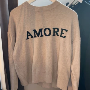 Beige tröja med texten 'AMORE' - Säljer en beige tröja med texten 'AMORE' i svart på framsidan. Tröjan har en rund hals och långa ärmar.