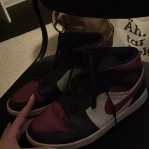 Nike Air Jordans 1 i svart, röd och vit - Snygga Nike Air Jordans i klassisk färgkombination av svart, röd och vit. Skorna har snörning och en ikonisk design med en röd sula. Perfekta för dig som vill ha en stilren och sportig look. Kan sänka priset vid snabb affär