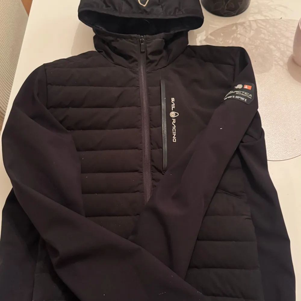 Snygg svart jacka från Sail Racing med dragkedja och huva. Jackan har en stilren design med en bröstficka och logotyp på ärmen. Perfekt för kyligare dagar. Bra skick! Det som syns på bilden är så den ser ut!. Takit.
