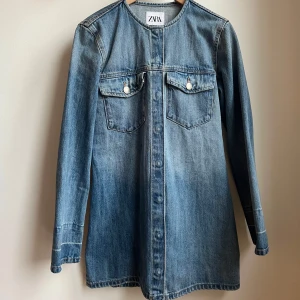 Jeansklänning från Zara - Snygg jeansklänning från Zara i en klassisk blå denim. Klänningen har långa ärmar och två bröstfickor med knappar. Perfekt för en avslappnad stil med en touch av elegans.