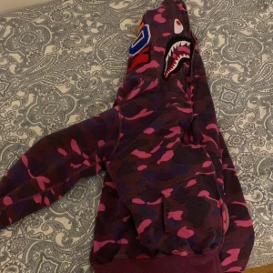 Camouflage hoodie från A Bathing Ape - Säljer en cool hoodie från A Bathing Ape med ett unikt rosa och lila camouflage-mönster. Hoodien har en dragkedja och en hajdesign på huvan med broderade detaljer. Perfekt för dig som vill sticka ut! Använd men gott skick