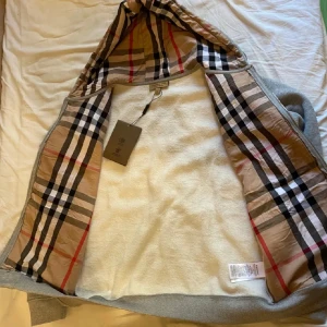  Burberry hoodie - Snygg grå huvtröja från Burberry med klassiskt rutigt foder i huvan. Tröjan har en broderad logotyp på bröstet och dragkedja framtill. Perfekt för en stilren och bekväm look.