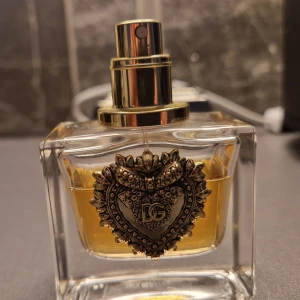 Dolce & Gabbana Devotion Eau de Parfum - Elegant parfymflaska från Dolce & Gabbana med en lyxig hjärtformad medaljong. Flaskan innehåller 30 ml av doften Devotion, en Eau de Parfum. Tillverkad i Italien, perfekt för den som söker en sofistikerad och unik doftupplevelse. Se bild för mängd som är kvar. Utan kork.
