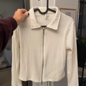 Vit ribbad tröja med dragkedja  - Säljer en stilren vit ribbad tröja från. Tröjan har en dragkedja framtill och långa ärmar, perfekt för lager-på-lager. Den har en klassisk krage och är i en croppad modell.