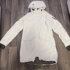 Vit parkajacka från Didriksons - Säljer en stilren vit parkajacka från Didriksons. Jackan har en praktisk huva och flera fickor framtill. Den är långärmad med justerbara ärmslut och dragkedja framtill. Perfekt för kyliga dagar.