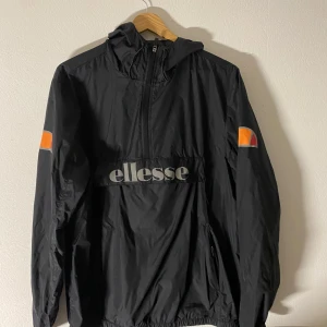 Svart vindjacka från Ellesse - Snygg svart vindjacka från Ellesse med dragkedja och logga på bröstet. Jackan har långa ärmar och en praktisk huva. Perfekt för blåsiga dagar. Storlek S.