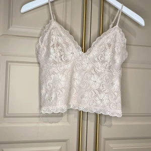 Beige spetslinne - Säljer ett vackert beige spetslinne med smala axelband och v-ringad design. Toppen har ett blommigt spetsmönster och är croppad i modellen. Perfekt för en somrig look!