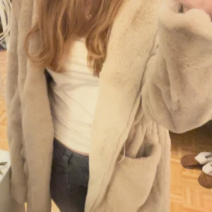 Beige pälsjacka - Mysig beige pälsjacka med långa ärmar och en avslappnad passform, det finns också snörning i midjan så man kan få den att sitta figurnära. Perfekt för kyliga dagar när du vill hålla dig varm. Den har också en dragkedja och stor luva. I nyskick.