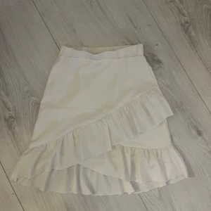 Vit omlottkjol med volang - Snygg vit omlottkjol med volangdetaljer. Perfekt för en somrig look. Kjolen har en omlott-design som ger en fin rörelse och stil.