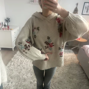 Beige stickad hoodie med blommönster - Mysig beige stickad hoodie med blommönster i rött, brunt och blått. Tröjan har en avslappnad passform och långa ärmar, perfekt för kyliga dagar. Den har en huva för extra värme och stil.
