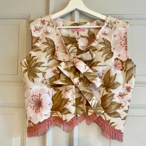 Blommig väst med fransar - Vest med blomsterprint og frynser. Vesten har en V-udskæring og lukkes med to sløjfe foran.  Den er syet af upcyclet vintage tekstil   Onesize ˜ brystmål: 110  Tags: #slowmade #slowfashion #hawaii #hawaiiblomst #vest #sløjfe #handmade #vintagetextile #vintage #håndsyet #upcycle #redesign #smallbizz 