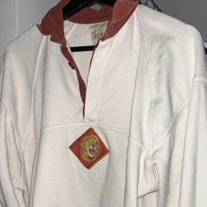 Vit tröja med lejonmotiv från Edison - Säljer en vit tröja från Edison med ett coolt lejonmotiv på framsidan. Tröjan har en mörk röd krage och knappdetalj. Perfekt för en avslappnad stil. Pris kan diskuteras