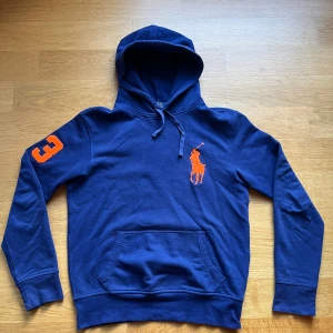 Blå hoodie från Ralph Lauren - Säljer en snygg blå hoodie från Ralph Lauren med en stor orange logga på bröstet och siffran 3 på ärmen. Tröjan har en klassisk känguruficka och justerbar huva med snören. Perfekt för en avslappnad stil.