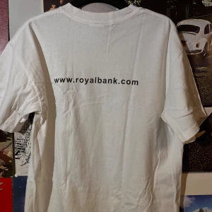 Vit t-shirt från Gildan - Vit t-shirt från Gildan med tryck på både fram- och baksida. Framsidan har en liten logotyp med texten 'Bank On-Line' och 'Canada'. Baksidan har en webbadress. Tillverkad i kraftig bomull, perfekt för en avslappnad stil.