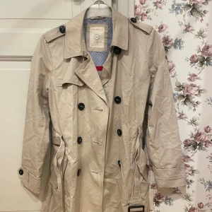 Beige trenchcoat från Esprit - Snygg beige trenchcoat från Esprit med klassisk design. Jackan har knappar och bälte i midjan för en stilren look. Perfekt för vår och höst.