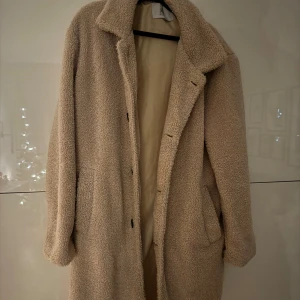 Beige teddykappa från Sixth June - Mysig beige teddykappa från Sixth June. Jackan har en lång och rak passform med knappar framtill och två sidofickor. Perfekt för kyliga dagar med sin mjuka och varma textur.