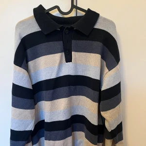 Randig pikétröja från Redgreen - Säljer en randig pikétröja från Redgreen i svart, grått och beige. Tröjan har en klassisk krage med knappar och långa ärmar. Perfekt för en avslappnad stil.