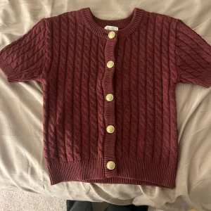 Vinröd kabelstickad kofta med korta ärmar - Säljer en snygg och populär vinröd kabelstickad kofta med korta ärmar och dekorativa knappar framtill. Perfekt till allt. Sparsamt använd.