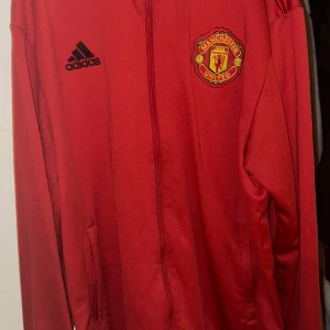 Röd Manchester United jacka från Adidas - Säljer en röd jacka från Adidas med Manchester United-logga på bröstet. Jackan har dragkedja och långa ärmar, perfekt för fotbollsfans. Den är stilren och sportig med Adidas-logga i svart.