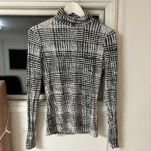 Svartvit mönstrad polotröja från H&M - Snygg svartvit polotröja från H&M med ett unikt rutigt mönster. Tröjan har långa ärmar och en tight passform, perfekt för en stilren look. Passar bra till både jeans och kjol.