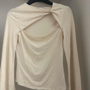 Beige långärmad topp med twistdetalj - Säljer en stilren beige långärmad topp med unik twistdetalj framtill. Perfekt för att ge en extra touch till din outfit. Toppen har en normal passform och är tillverkad i ett mjukt material.