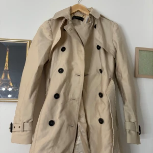 Beige trenchcoat med knappar - Snygg beige trenchcoat med klassiska svarta knappar och bälte i midjan. Perfekt för höst och vår. Långärmad design med stilrena detaljer.