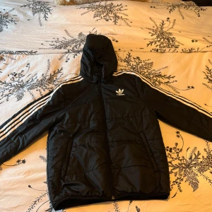 Svart dunjacka från Adidas - Säljer en svart dunjacka från Adidas med huva och de klassiska tre ränderna längs ärmarna. Jackan har dragkedja och är perfekt för kyligare väder. Tillverkad av Primegreen-material för en hållbar touch.