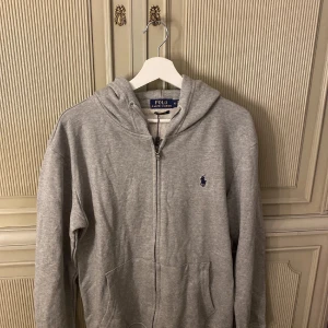 Grå hoodie från Ralph Lauren - Säljer en klassisk grå hoodie från Ralph Lauren med dragkedja och huva. Den har en broderad logga på bröstet och är perfekt för en avslappnad stil. Passar bra till både jeans och joggers. Tröjan är i nyskick och har inga tecken på defekter eller liknande. Priset kan diskuteras vid snabb affär😁