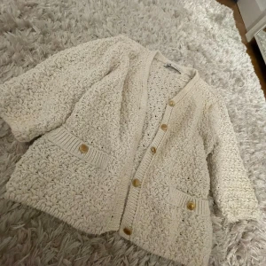 Beige stickad kofta med knappar - Mysig beige stickad kofta med stora knappar och två fickor framtill. Perfekt för kyliga dagar och ger en avslappnad look. Koftan har en lös passform och trekvartsärmar.