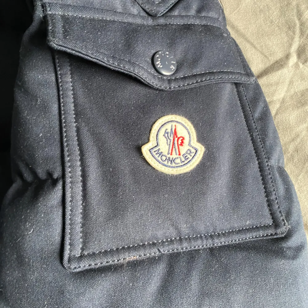 Säljer en snygg mörkblå dunjacka från Moncler med huva och dragkedja. Jackan har långa ärmar och praktiska fickor med knappar. Perfekt för kyliga dagar.. Takit.