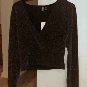 Svart glittrig topp från Vera Moda  - Snygg svart glittrig topp från Vera Moda med v-ringning och långa ärmar. Perfekt för en kväll ute eller festliga tillfällen💕