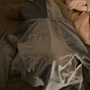 Grå velour hoodie från Juicy Couture - Säljer en mjuk och bekväm grå velour hoodie från Juicy Couture med glittrande stenar på ryggen. Perfekt för en avslappnad stil med en touch av glamour. Hör av dig vid intresse! 