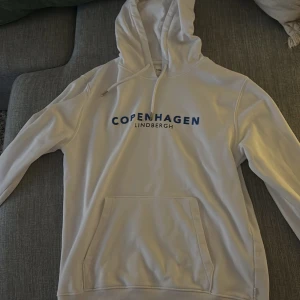 Vit hoodie från Lindbergh - Säljer en stilren vit hoodie från Lindbergh med texten 'Copenhagen' i blått på bröstet. Den har en klassisk känguruficka och justerbar dragsko i huvan. Perfekt för en avslappnad stil. Se bild 2 så är det en prick som ej går bort