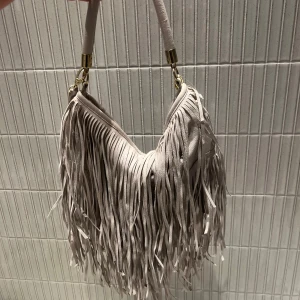 Beige axelväska med fransar - Snygg beige axelväska med fransar och gulddetaljer. Perfekt för att ge din outfit en bohemisk touch. Väskan har en bekväm axelrem och är tillverkad i ett mjukt material.