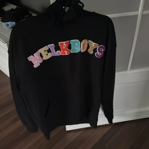 Svart hoodie från Nelk Boys - Säljer en svart hoodie från Full Send med färgglada bokstäver som bildar texten 'NELKBOYS' på framsidan. Hoodien har en klassisk design med långa ärmar och en bekväm huva. Perfekt för en avslappnad stil.