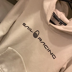 Vit hoodie från Sail Racing - Säljer en stilren vit hoodie från Sail Racing med svart logotyp på bröstet. Den har en klassisk känguruficka och en bekväm huva. Perfekt för en avslappnad stil.