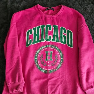 Rosa sweatshirt med Chicago-tryck - Säljer en cool rosa sweatshirt med stort grönt 'CHICAGO' tryck på framsidan. Tröjan har långa ärmar och rund halsringning. Perfekt för en avslappnad stil.