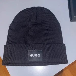 Svart mössa från Hugo boss - Snygg svart mössa från Hugo i ribbad design. Perfekt för att hålla värmen under kyliga dagar. Mössan har en stilren etikett framtill med märkesnamnet.