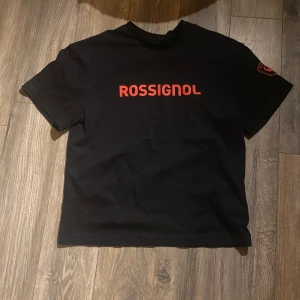 Svart t-shirt från Rossignol - Väldigt sällsynt!!!svart t-shirt från Rossignol i 100% ekologisk bomull. T-shirten har korta ärmar och ett rött tryck med Rossignol-loggan på bröstet och ärmen. Perfekt för en casual look. 