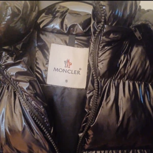 Svart dunjacka från Moncler - Snygg svart dunjacka från Moncler med glansig finish. Jackan har en dragkedja framtill och praktiska fickor. Perfekt för kyliga dagar med sin varma och puffiga design. Huvan ger extra skydd mot väder och vind.