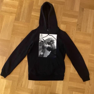 Tupac hoddie - Säljer en svart hoodie med ett stort Tupac-tryck på framsidan. Tröjan har en klassisk design med huva och långa ärmar. Perfekt för en avslappnad stil. Den är i fint skick och har inga defekter.
