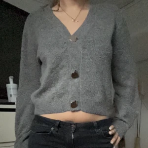 Grå kofta med knappar - Säljer en mysig grå kofta med stora knappar framtill. Koftan har en v-ringad design och långa ärmar, perfekt för kyligare dagar. Den är croppad och ger en trendig look. Det står M på pappen men passar mig bra som har S