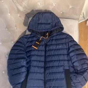 Blå pufferjacka från BOSS - Säljer en snygg blå pufferjacka från BOSS med huva och dragkedja. Jackan har ett quiltat mönster och är perfekt för kyligare dagar. Den har en stilren design med märkets logga på ärmen. Jackan har aldrig använts och om ni vill ha fler bilder de bara att skriva. Det är storlek S men passar också M